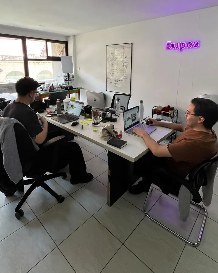 Oficina del equipo de Azenza: dos personas trabajando en sus computadoras, con el cartel de neón de Dupes en la pared.